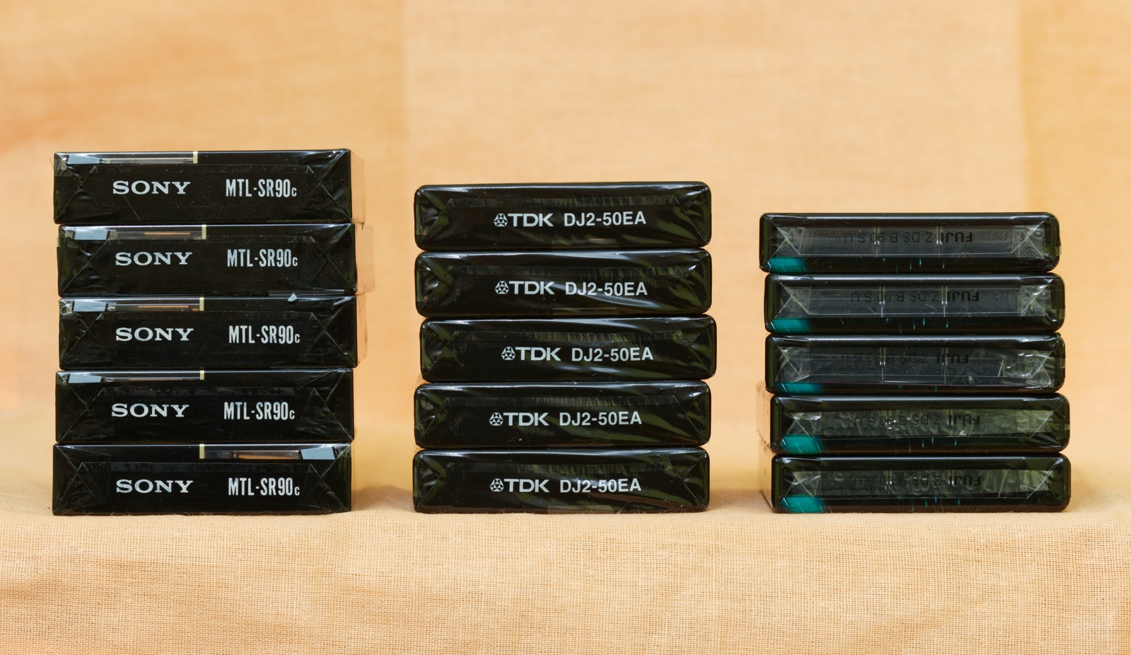 Three stacks of shrink-wrapped blank audio cassettes, Sony, TDK and Fuji, lined up on an orange cloth