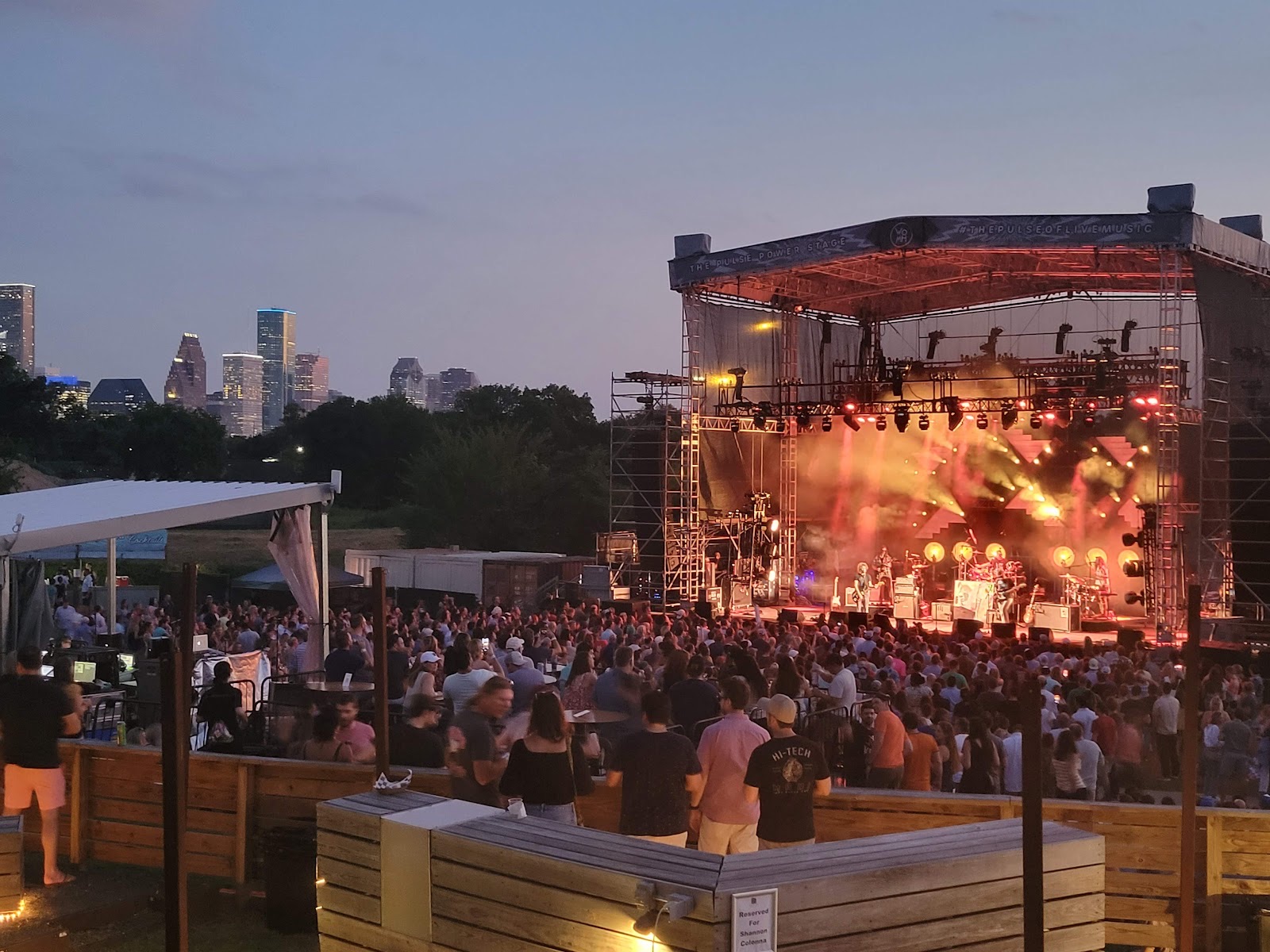 A crowd on the lawn at White Oak Music Hall watching a band on a lit outdoor stage at dusk, with the downtown Houston skyline behind the trees
