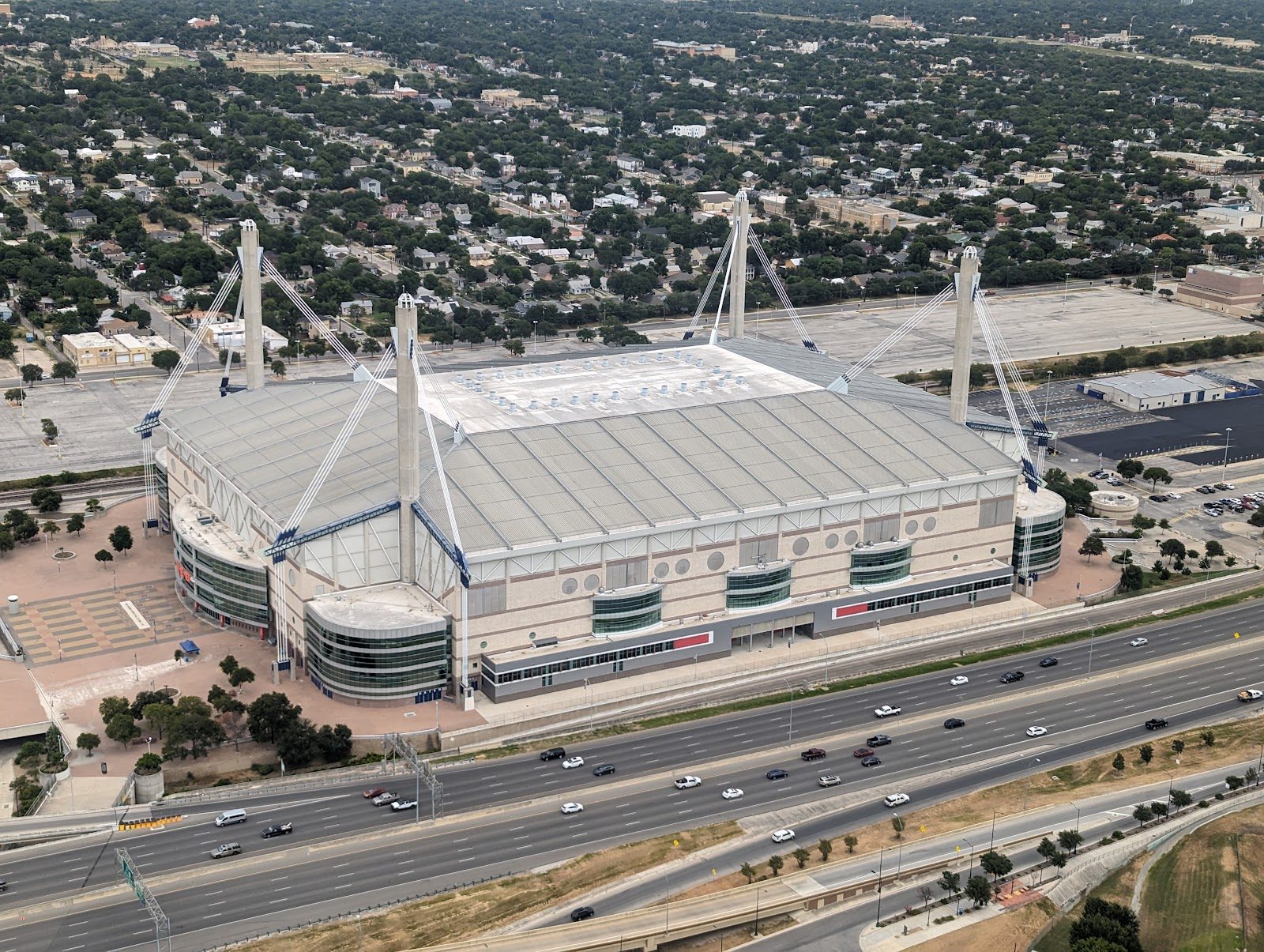 Aerial view of the Alamodome in San Antonio: a pale rectangular stadium with a flat white roof held by four tall concrete masts and cables, beside a busy freeway