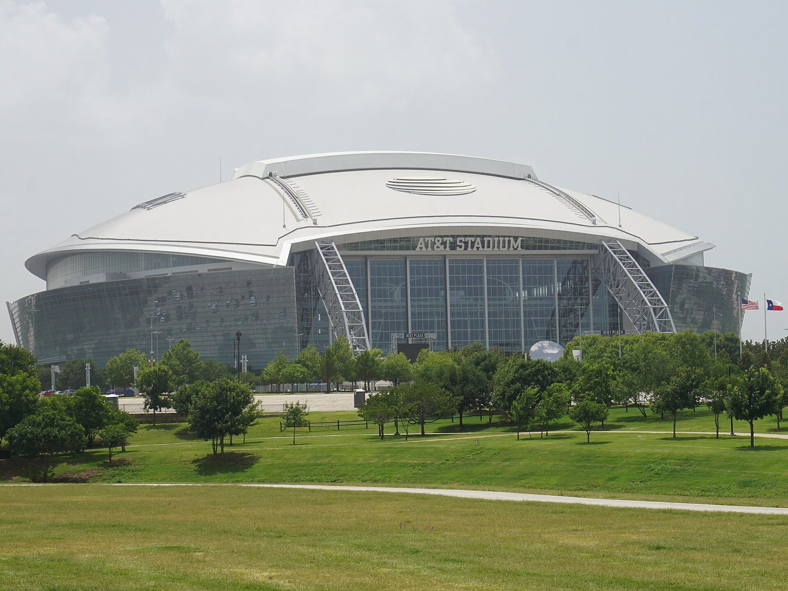 AT&T Stadium in Arlington seen across a green park: a white domed roof over a curved glass facade, with AT&T Stadium lettered above the tall glass entrance wall