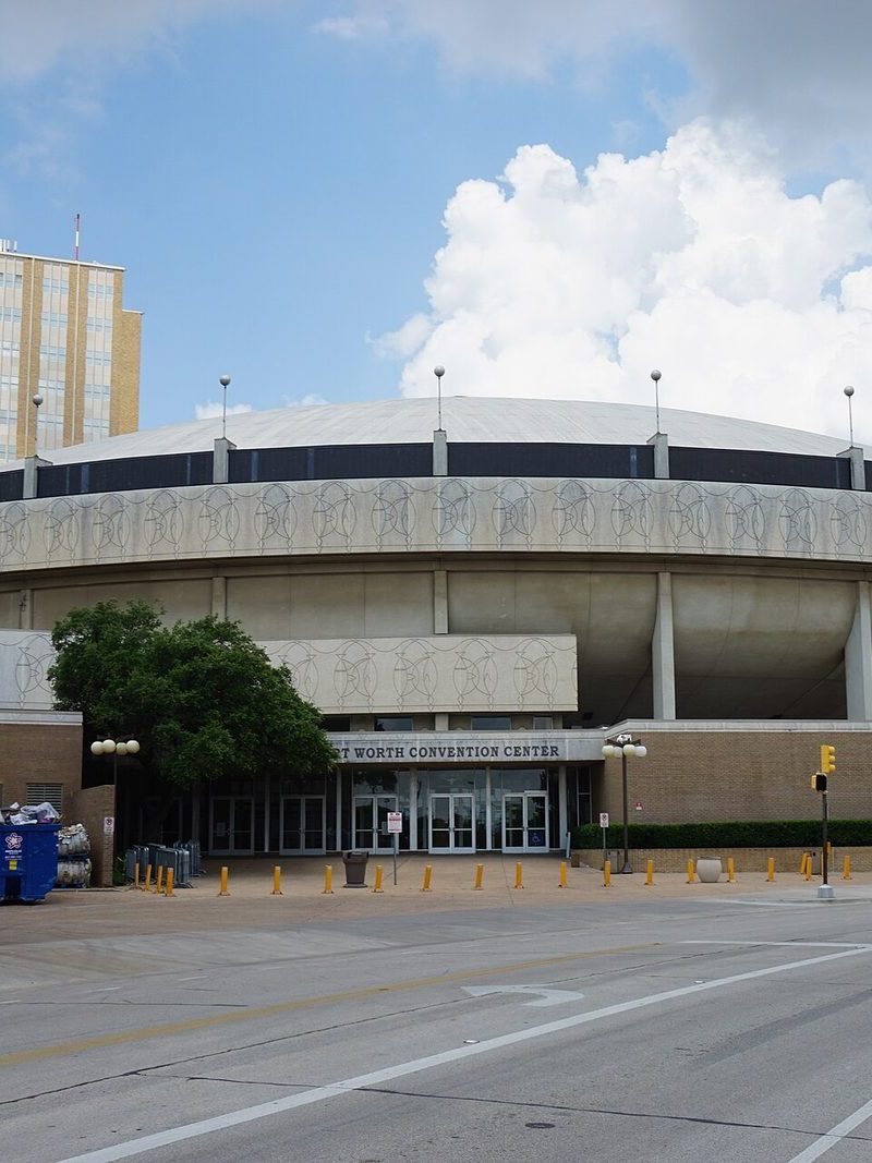 The round, saucer-shaped Fort Worth Convention Center arena under a cloudy sky, its patterned concrete band above glass entrance doors marked Fort Worth Convention Center