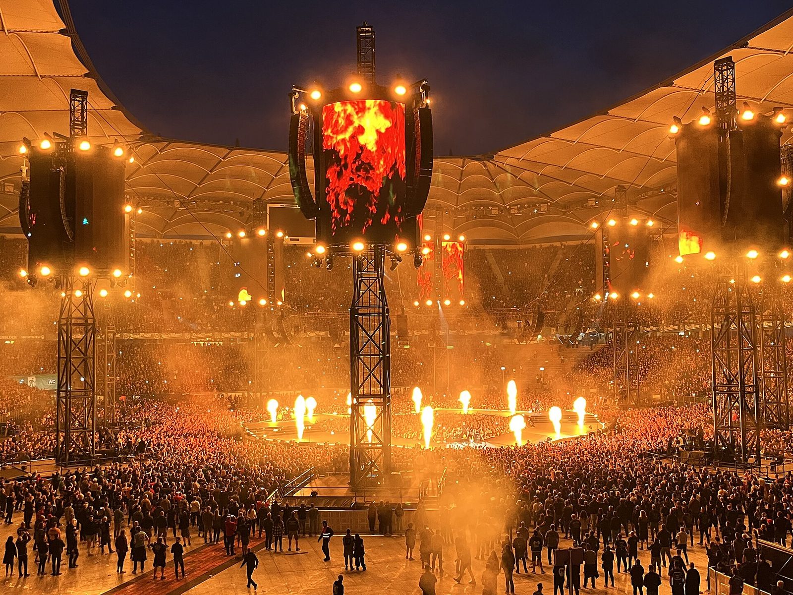 Metallica's M72 in-the-round stage at night, ringed by fans on the stadium floor, with jets of fire around the stage and flame graphics on a tall video tower in the middle