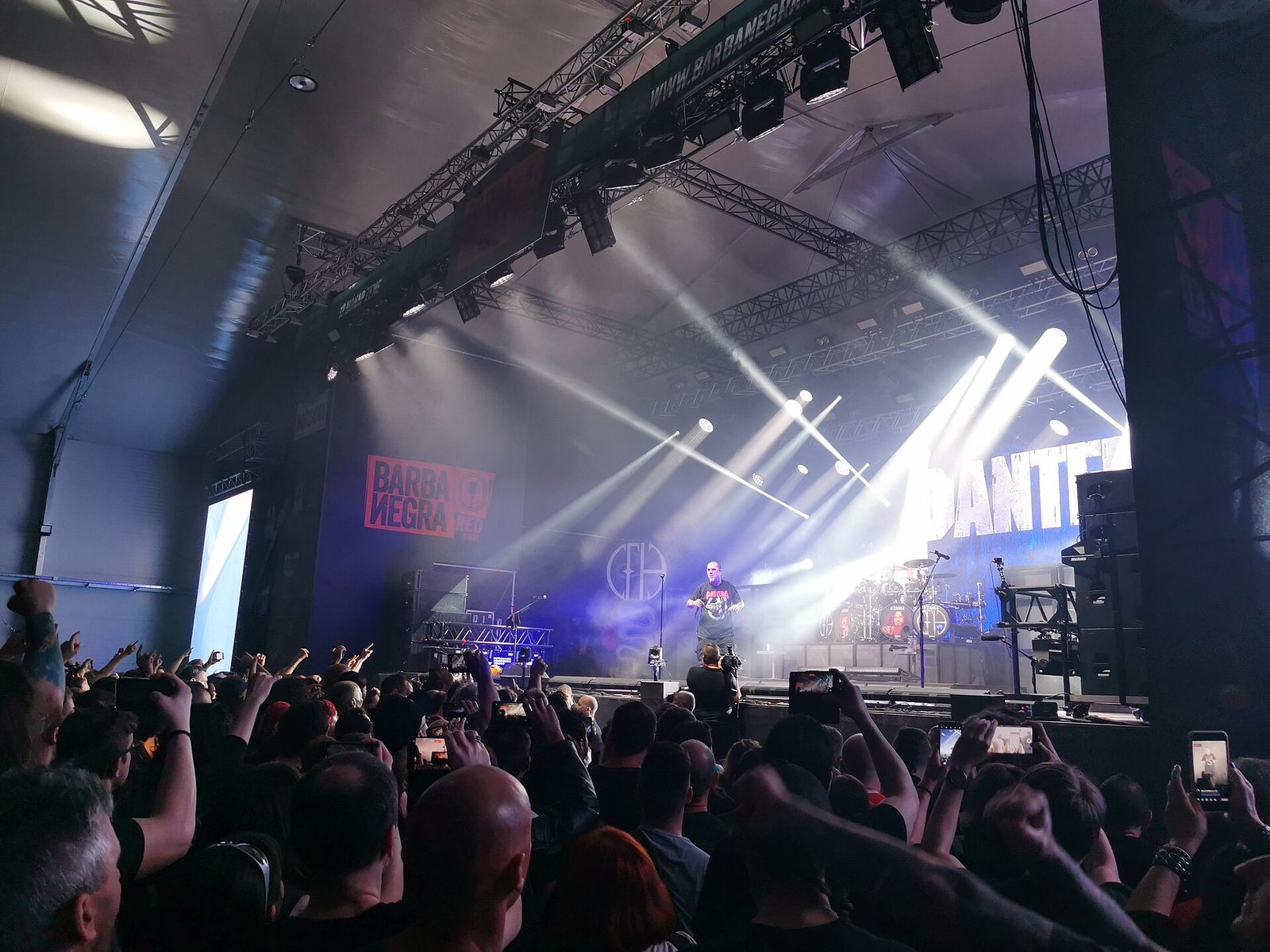 Phil Anselmo singing at the front of a smoky stage under white spotlights, with the Pantera logo on the backdrop and a crowd holding up phones in the foreground