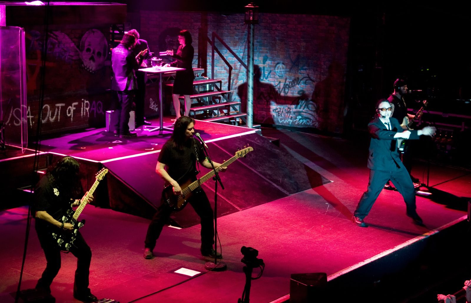 Queensrÿche on a pink-lit stage set built like a graffiti-covered alley: a guitarist and bassist at the front, a singer in a dark suit and sunglasses on the right, and actors at a table on a raised platform behind