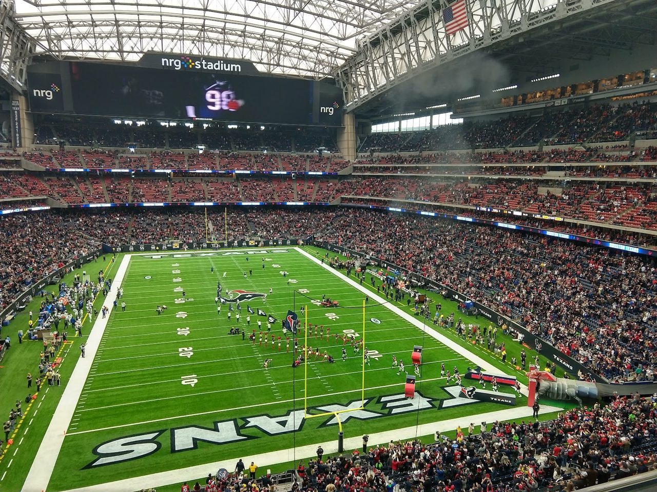 Inside the Houston stadium during a Texans game, NRG signage still up: packed red and blue stands around a green field with TEXANS painted in the end zone, under a closed roof of steel trusses