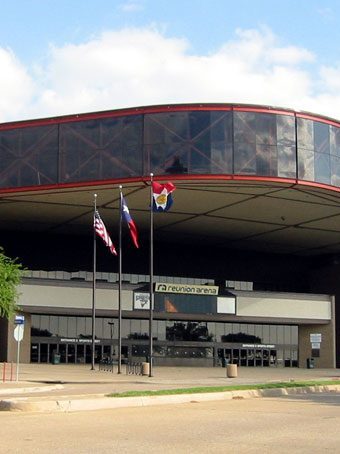 Reunion Arena's entrance in Dallas: a glass band with a red trim line wrapping the roof edge over a recessed lobby, with flags on poles out front