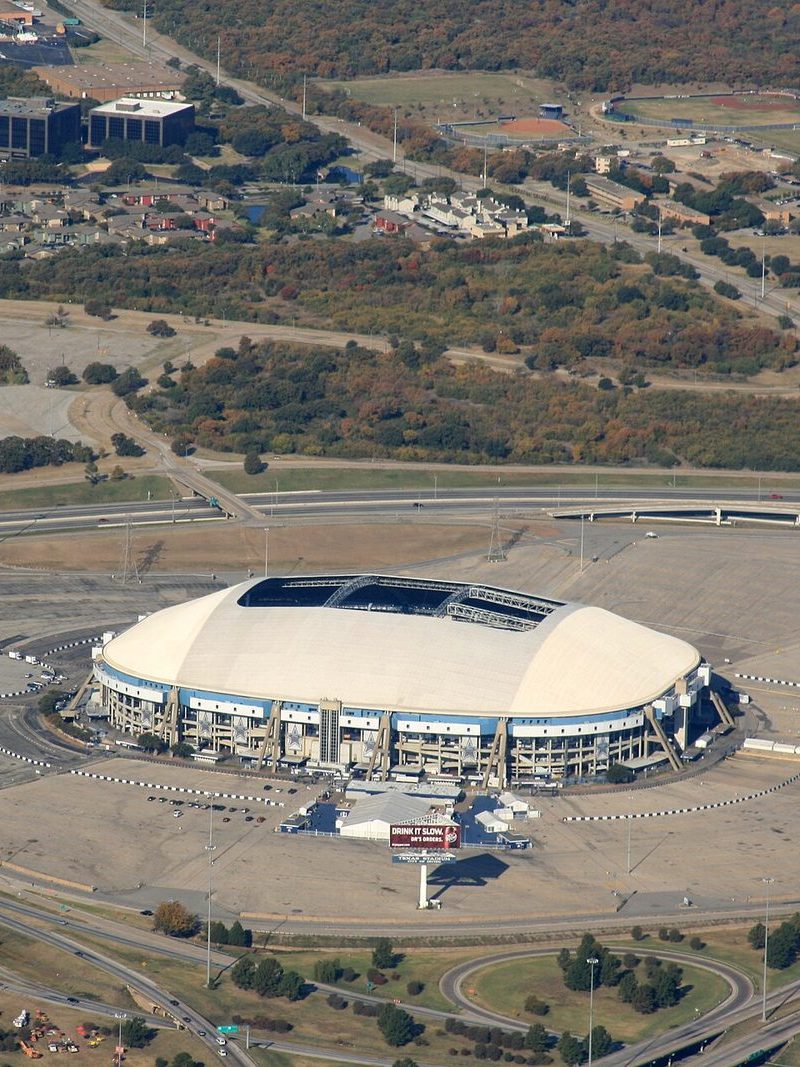 Aerial view of Texas Stadium in Irving, a white oval with an open hole in its roof, surrounded by empty parking lots and freeway interchanges