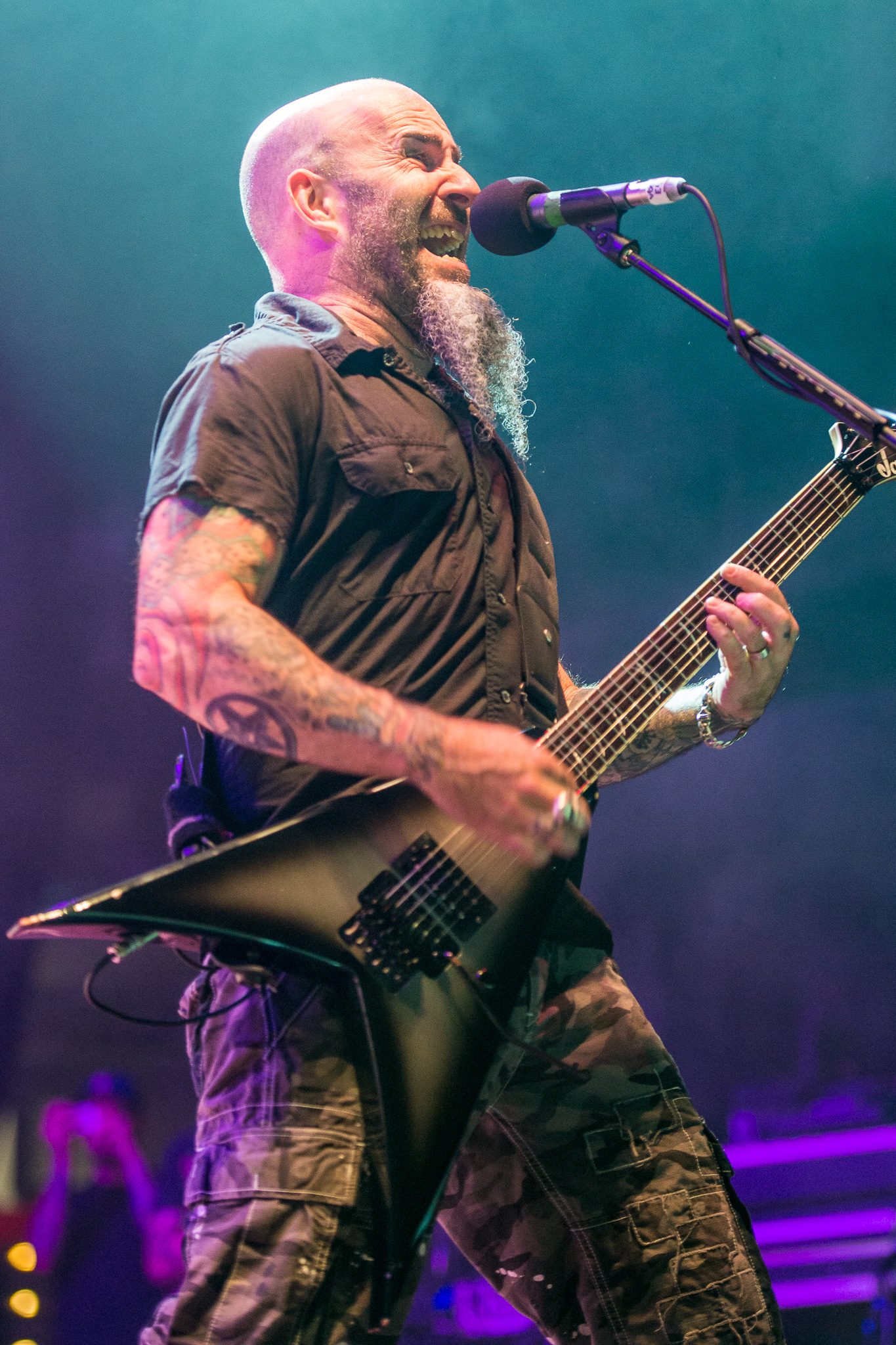 Scott Ian of Anthrax, bald with a long braided gray beard, shouting into a microphone while playing a black V-shaped guitar under blue and purple stage lights