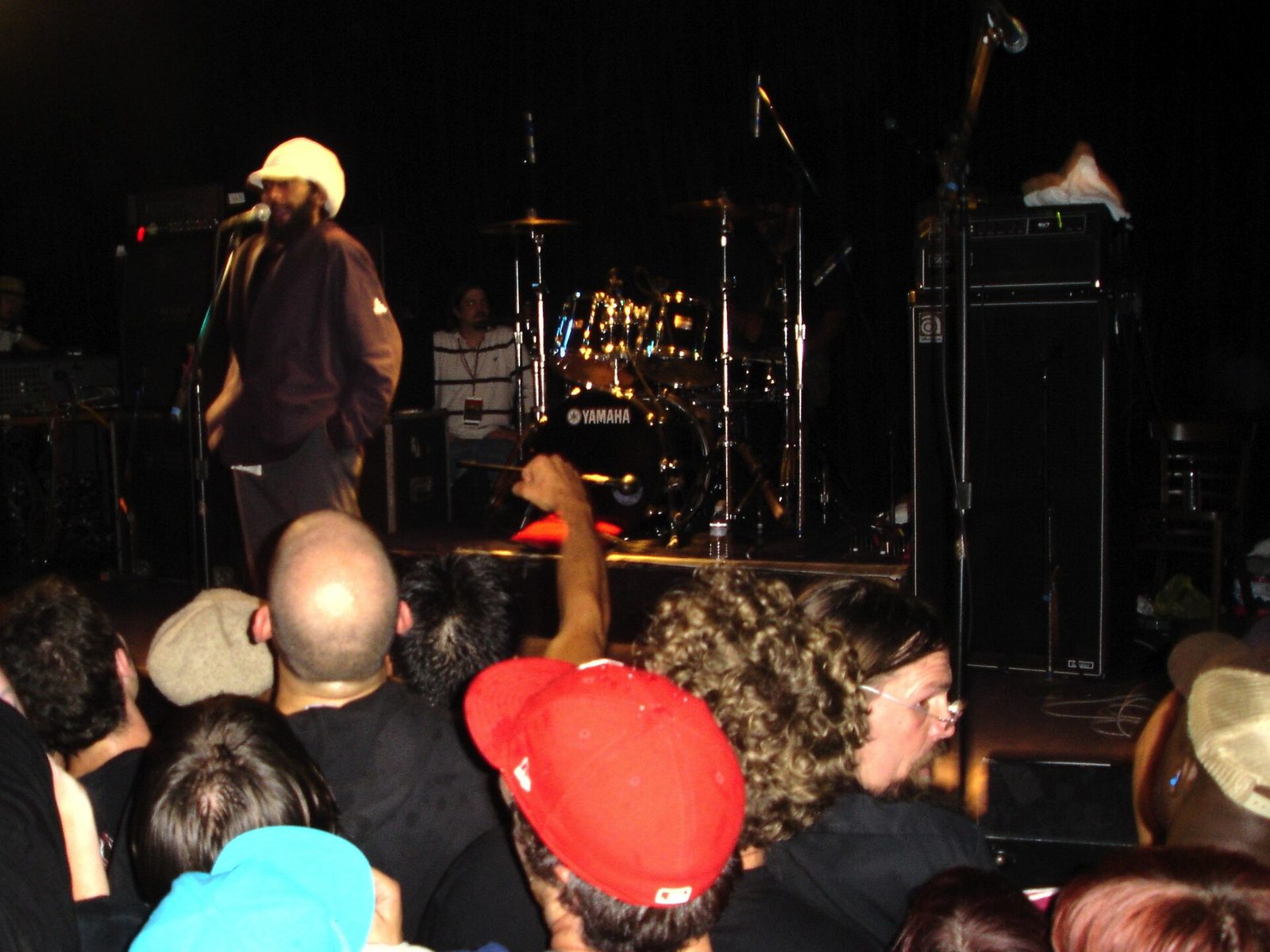 Bad Brains on a dark club stage, a singer in a white cap and brown jacket at the microphone and a drum kit behind him, the heads of the crowd filling the foreground