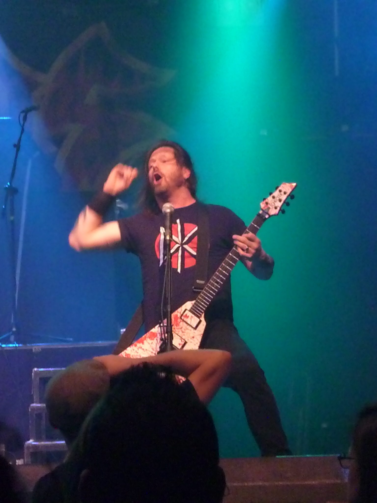 Gary Holt of Exodus, long-haired in a black T-shirt, shouting with his fist raised while playing a white blood-spattered V-shaped guitar under green and blue light, a fan's raised arms in the foreground