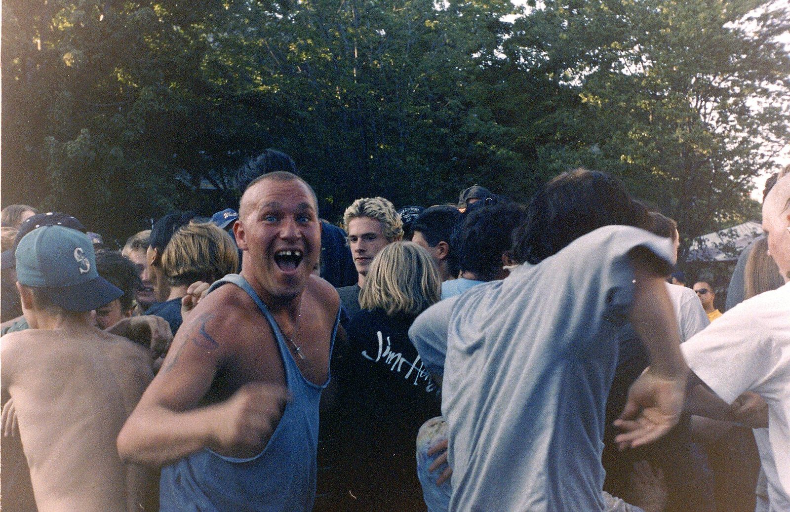 A shaven-headed young man in a gray tank top grins at the camera in the middle of an outdoor mosh pit, with shirtless and long-haired fans pushing around him under tall trees