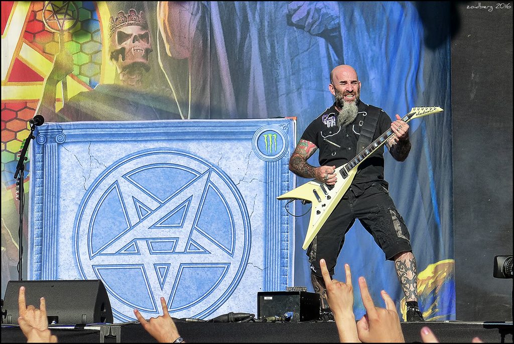 Anthrax on an outdoor festival stage, a bald, bearded guitarist with a yellow Flying V in front of a huge pentagram backdrop, fans' raised hands in the foreground