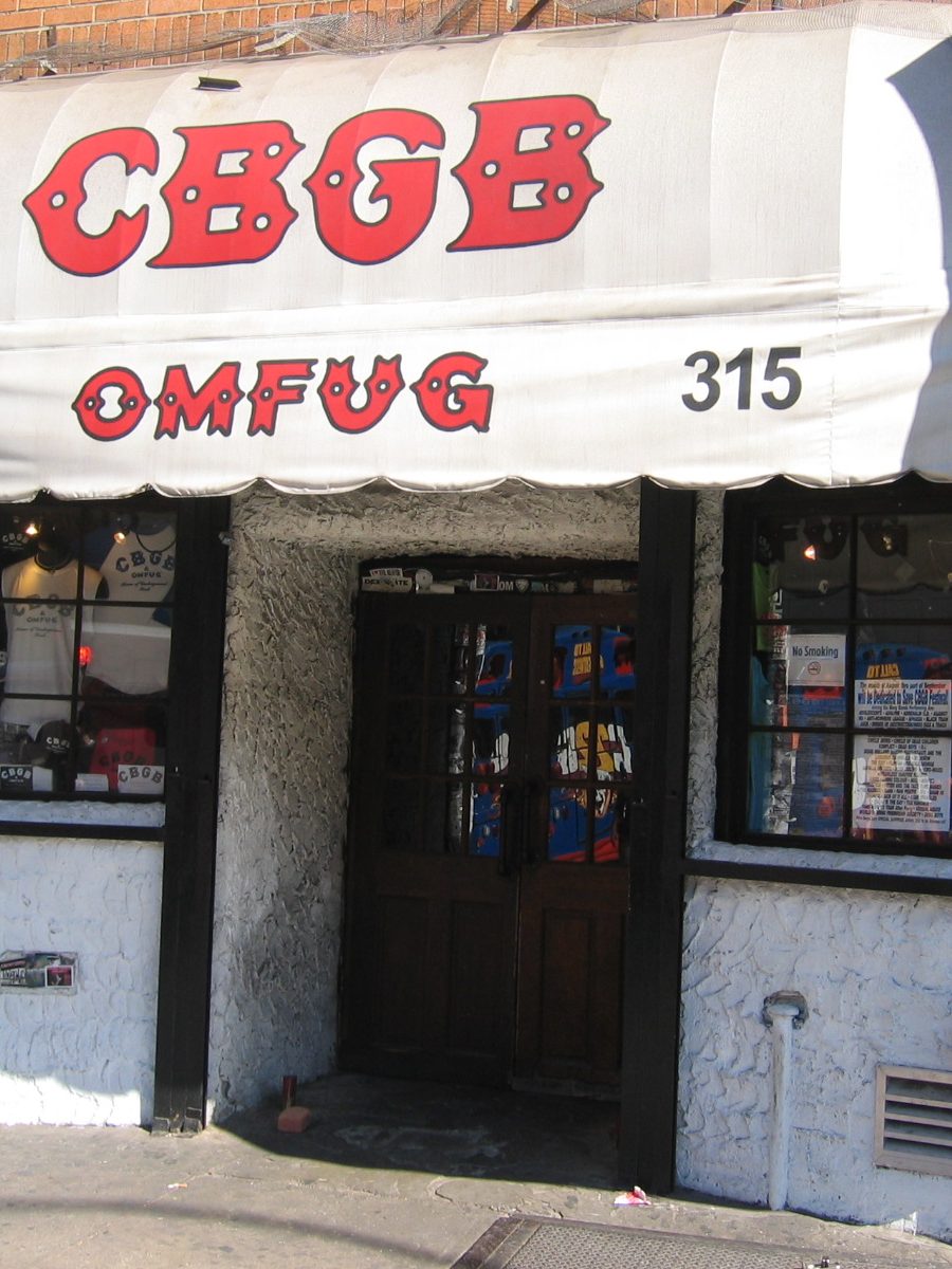 CBGB’s entrance at 315 Bowery: a white scalloped awning with red CBGB and OMFUG lettering and the number 315 over a dark wooden double door flanked by T-shirt-filled windows