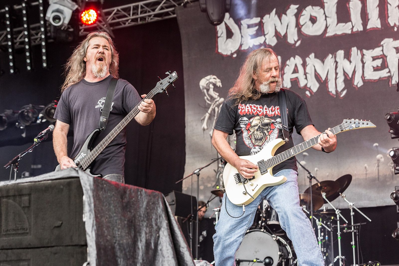 Two long-haired Demolition Hammer guitarists playing side by side on an outdoor stage in front of the band’s banner, a drummer behind them