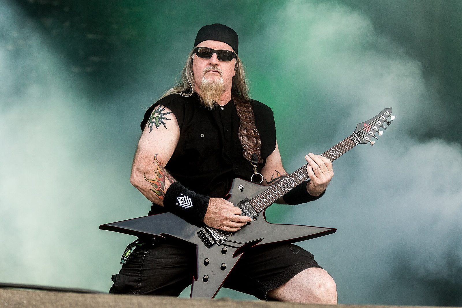 An Overkill guitarist in sunglasses and a black bandanna playing a black star-shaped guitar in front of drifting stage smoke