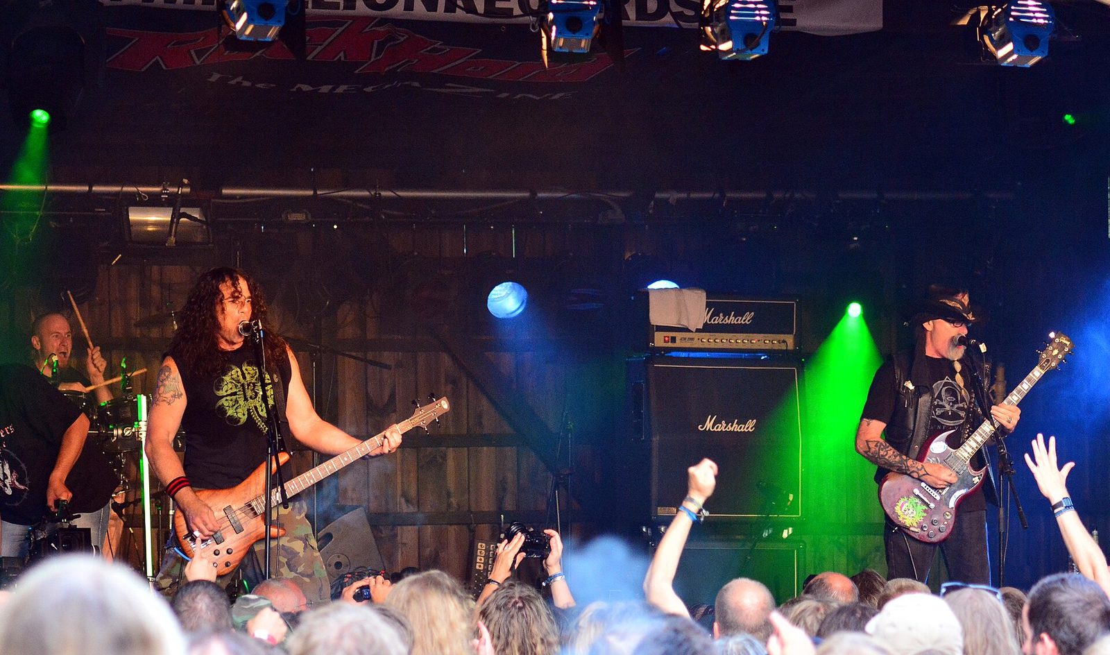 Whiplash on a small festival stage under green and blue lights, a guitarist singing at the microphone on the right and a bassist on the left, fans' arms raised in front
