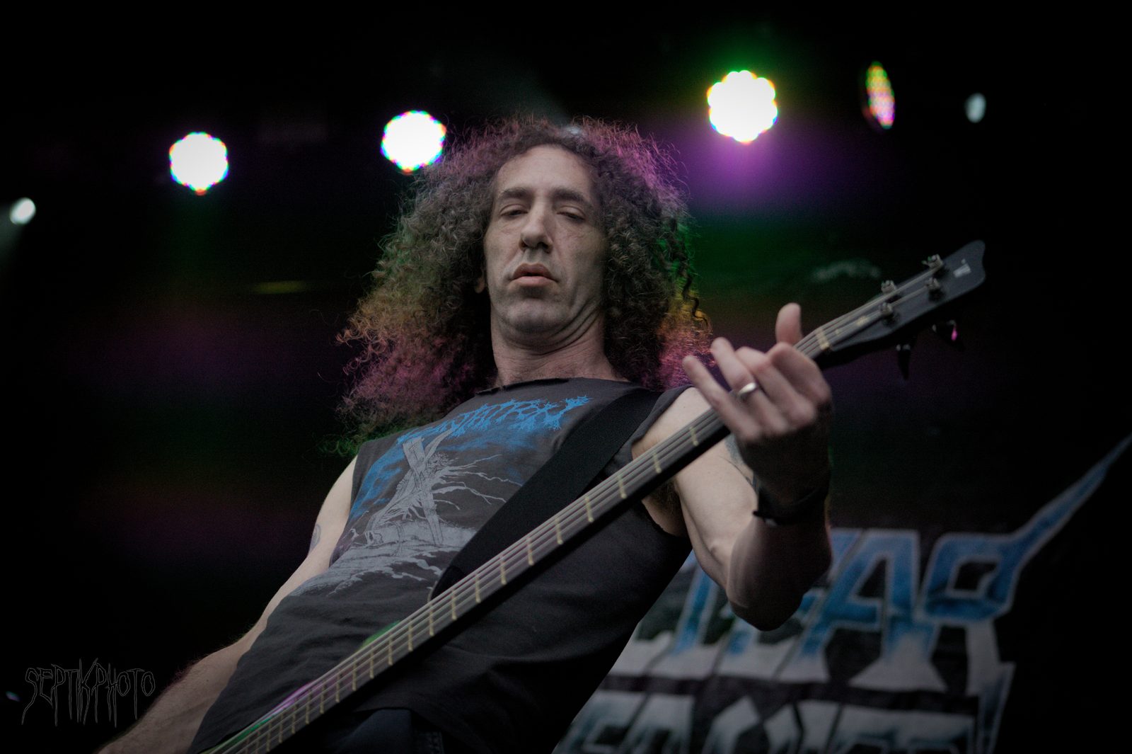 Nuclear Assault bassist Dan Lilker, long curly hair, playing a black bass under purple and white stage lights with the band's spiky logo banner behind him