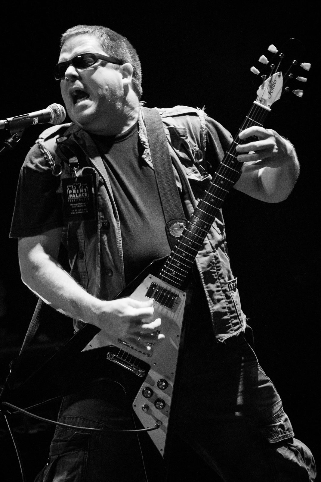 Black and white photo of John Connelly in sunglasses and a sleeveless vest, singing into a microphone while playing a Flying V guitar