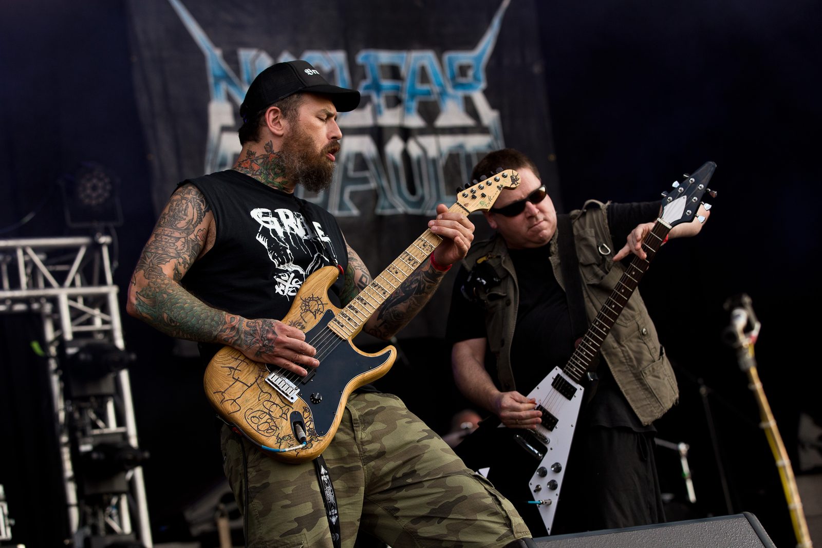 Two Nuclear Assault guitarists on an outdoor festival stage, a bearded, tattooed player with a natural-finish guitar in front and John Connelly with a Flying V behind him, the band's logo banner overhead