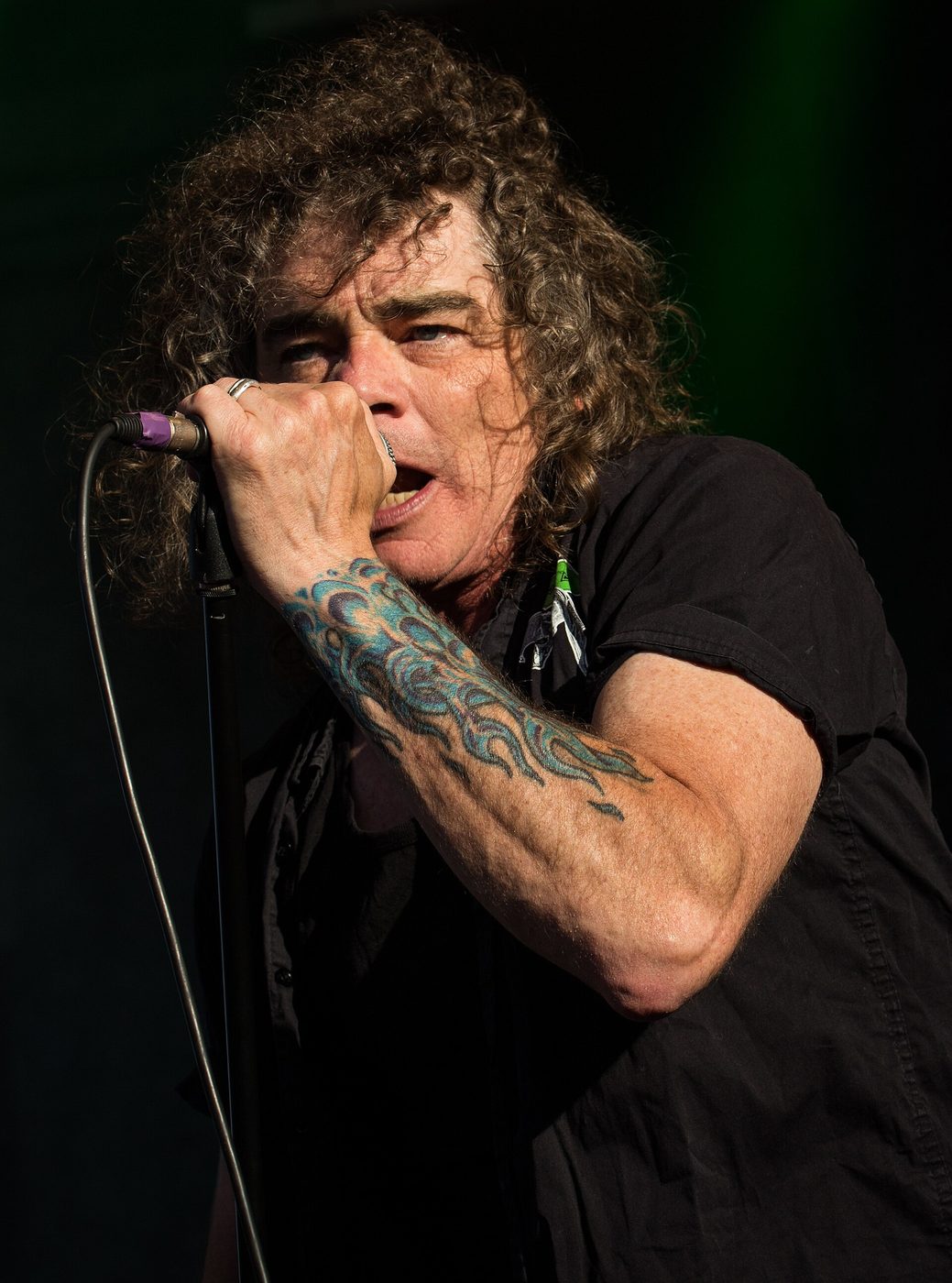 Bobby Blitz Ellsworth, curly hair and a black shirt, gripping a microphone close to his mouth, a blue flame tattoo on his forearm