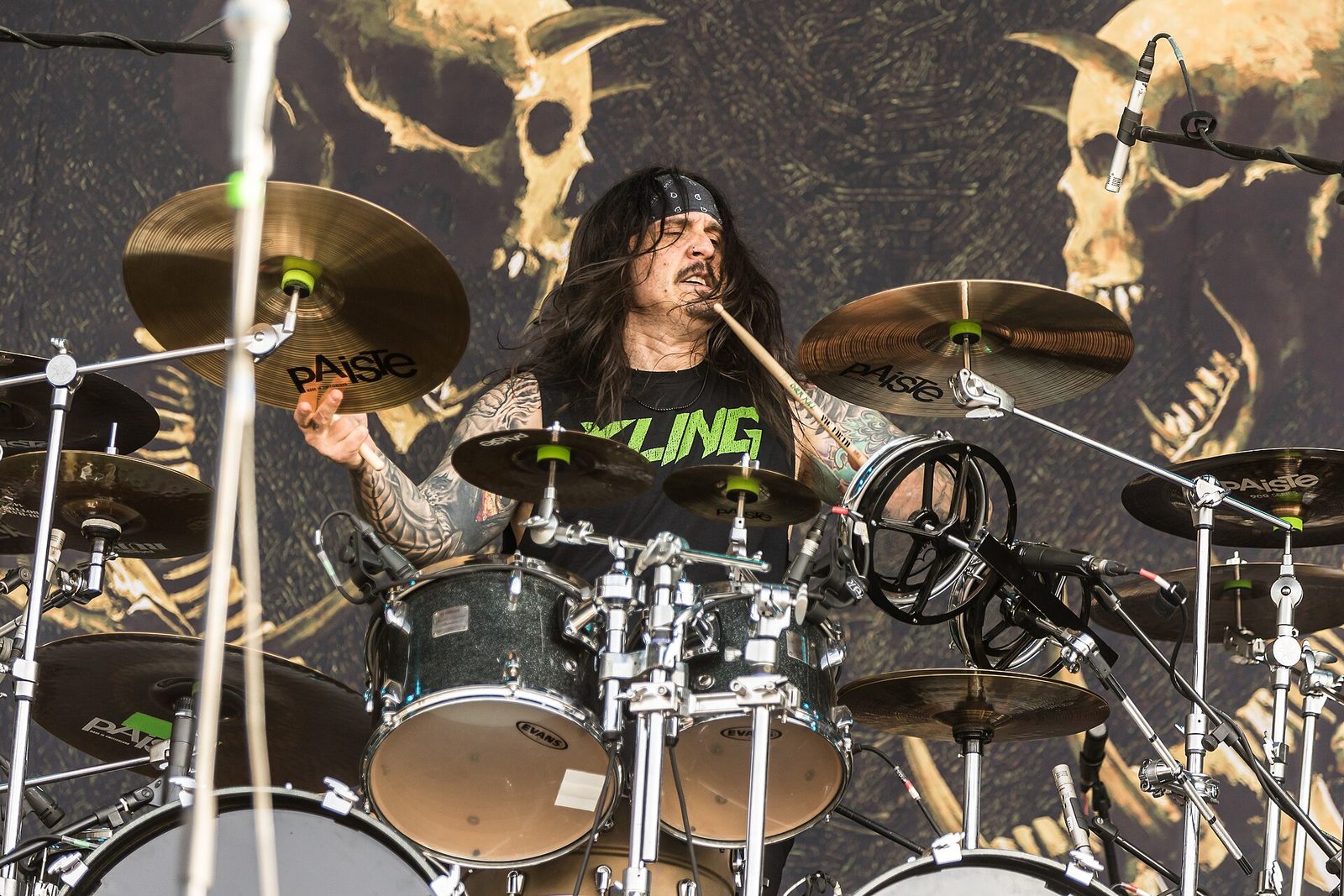 Drummer Jeramie Kling, long dark hair and a bandanna, mid-hit behind a kit of Paiste cymbals, wearing a black shirt with his name in green