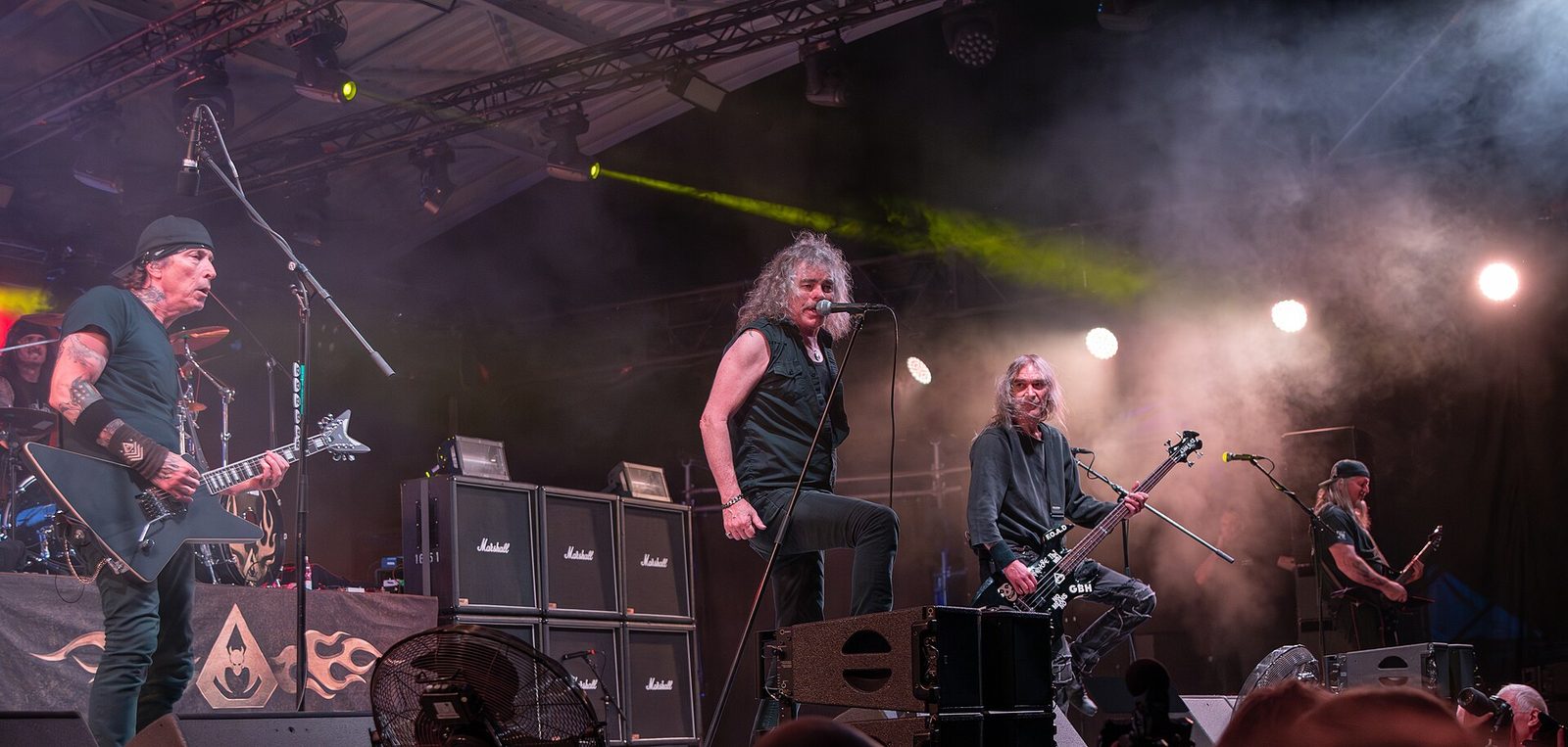 Overkill on a festival stage under a steel roof, Bobby Blitz Ellsworth singing at center between a guitarist with a black star-shaped guitar and a bassist, stage smoke and green lights behind them