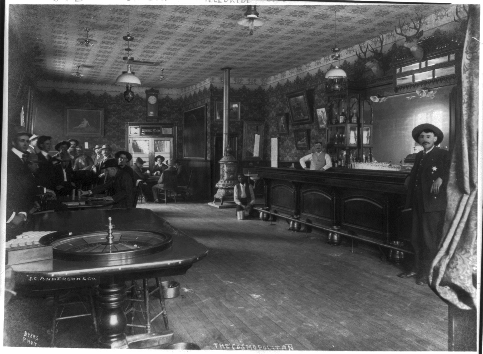1910 black and white photo of the Cosmopolitan Saloon in Telluride, Colorado, with a roulette table, gamblers at left, a long bar and a lawman with a star badge