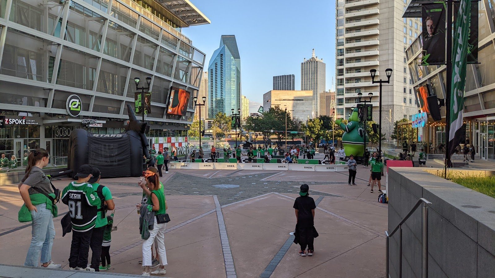 Dallas Stars fans in green jerseys gathered on the plaza outside the American Airlines Center before a game, with a street hockey rink and the downtown Dallas skyline behind