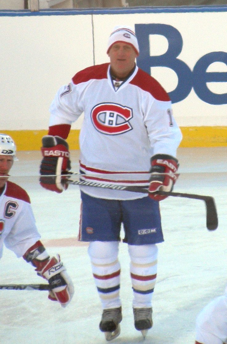 Craig Ludwig in a white Montreal Canadiens alumni jersey and white knit hat, holding his stick on an outdoor rink