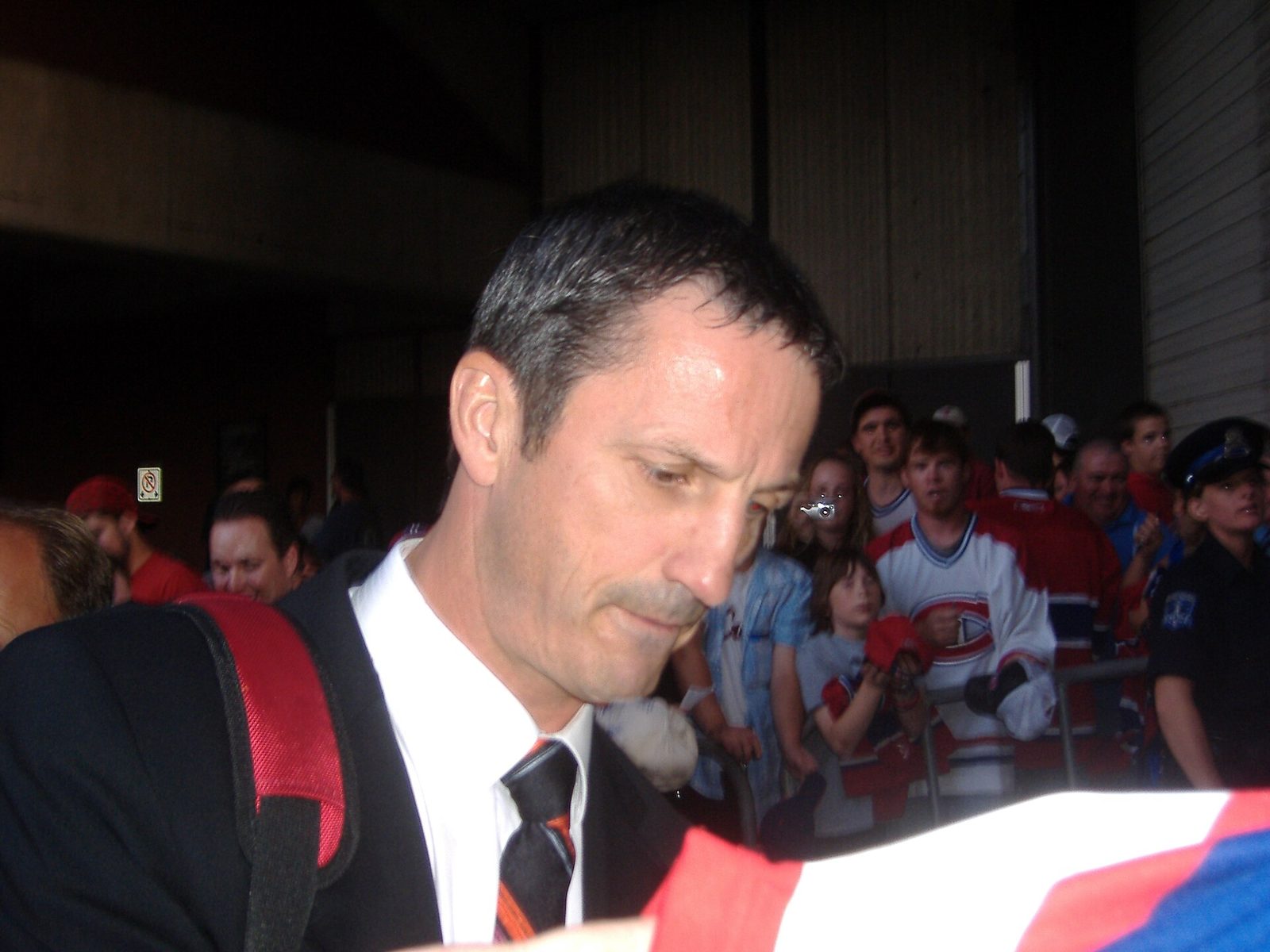 Guy Carbonneau in a suit and tie signing autographs outside an arena, with fans in Canadiens jerseys behind a barrier