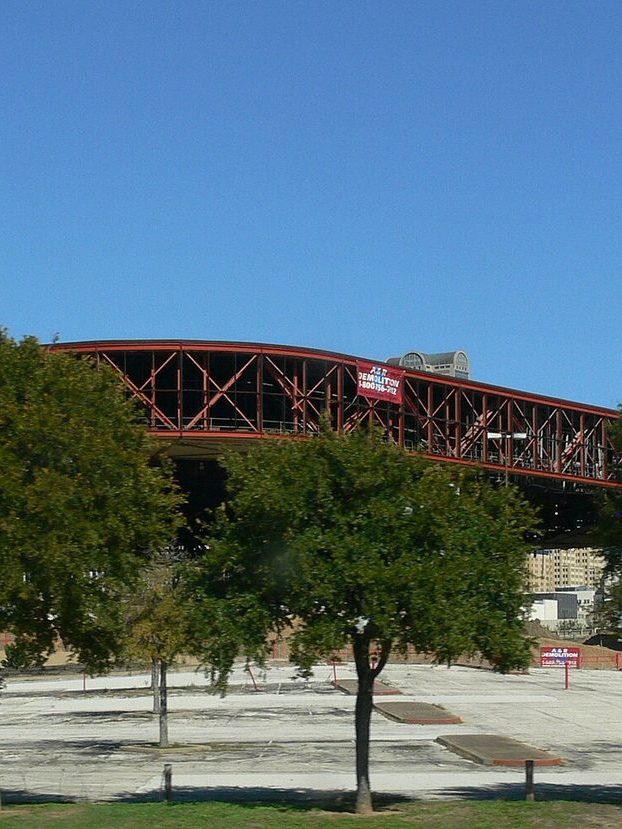 Reunion Arena in Dallas during demolition, its exposed red steel roof truss hung with a demolition company banner, above trees and an empty parking lot