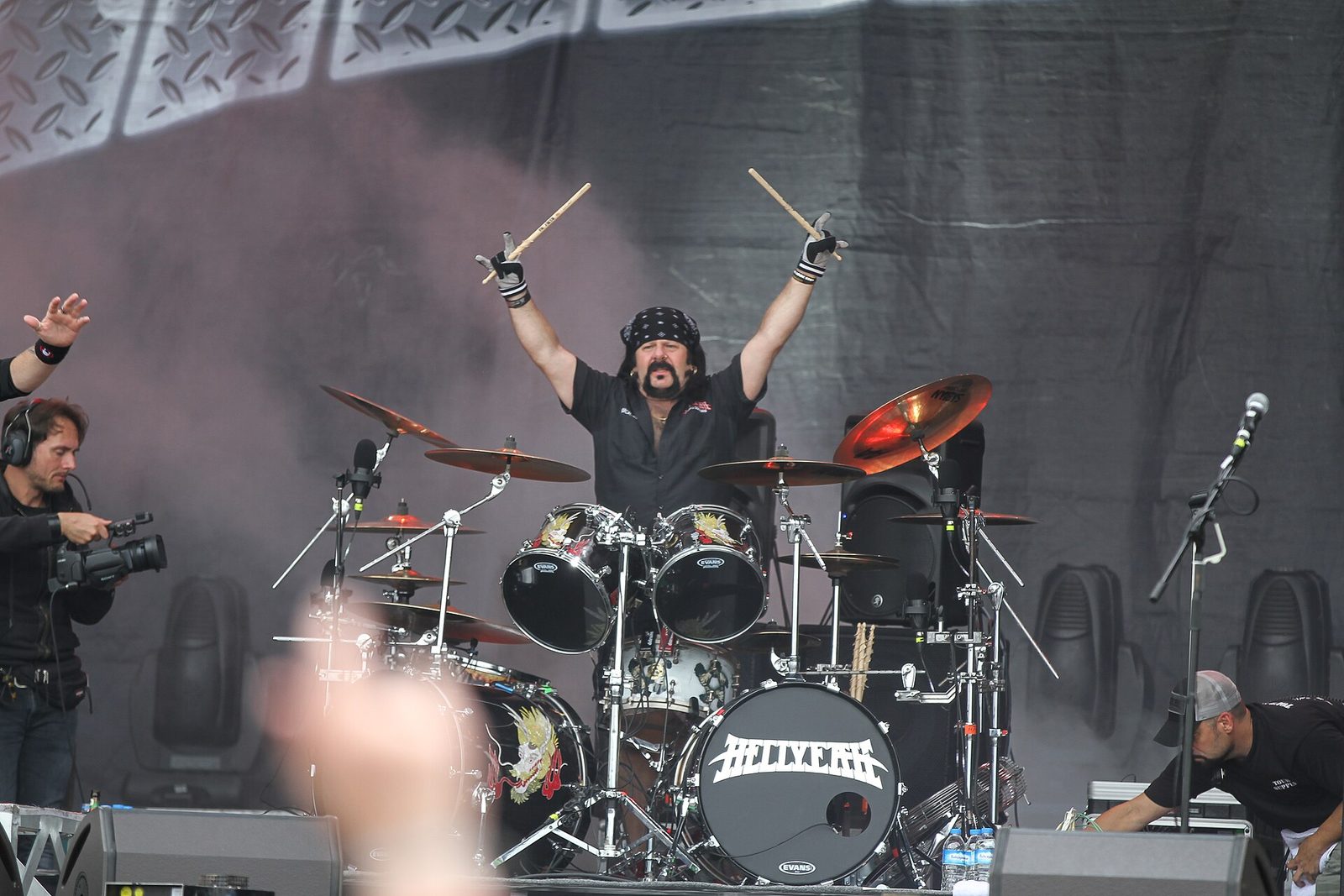 Vinnie Paul in a black bandana and black shirt raising both drumsticks over his head behind a drum kit with a Hellyeah bass drum head, at an outdoor festival stage