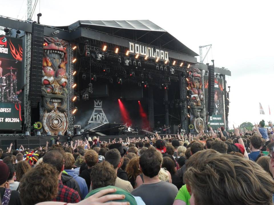 A packed crowd with raised hands in front of the Download Festival main stage at Donington Park, with giant carved totem figures on either side