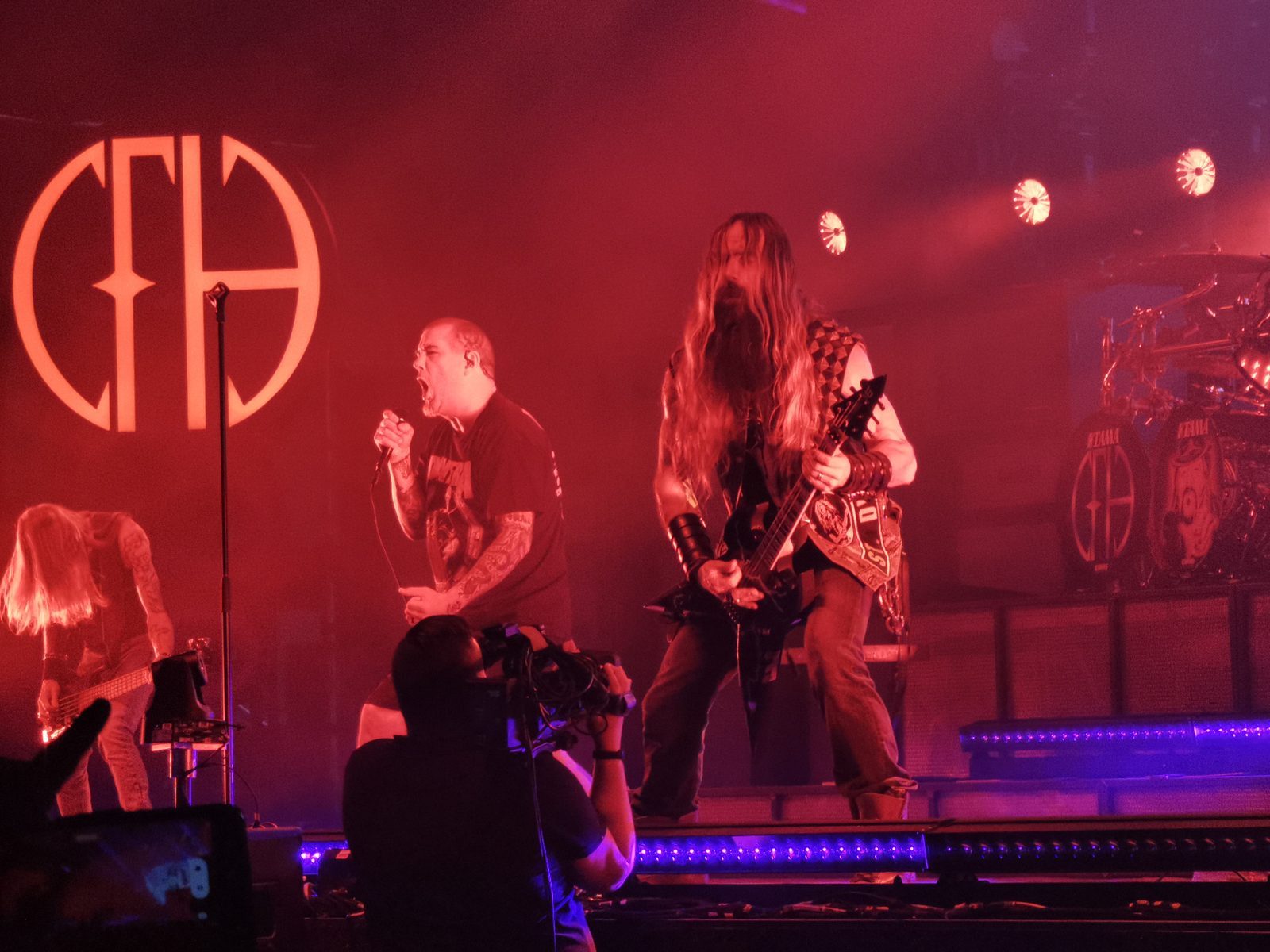 Phil Anselmo singing under red stage light beside a long-haired, bearded guitarist, with the Pantera PH logo on the wall and drum kit behind them