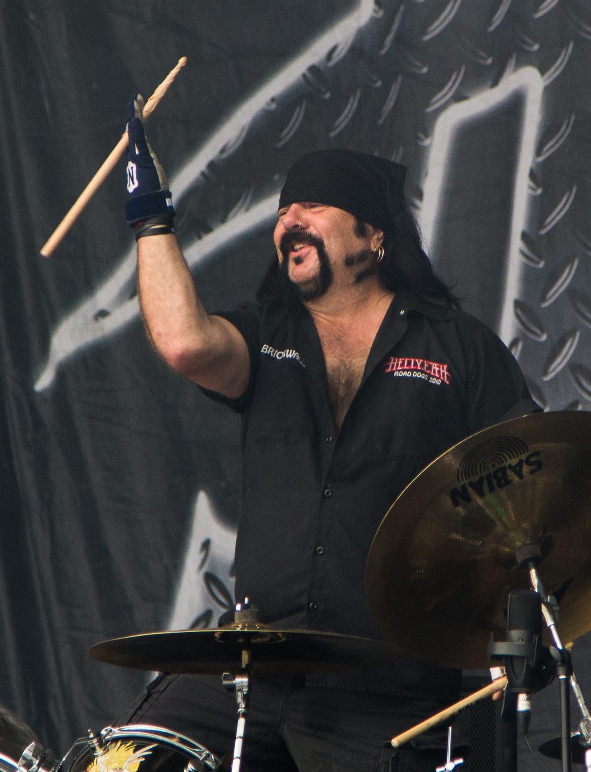 Vinnie Paul behind his drum kit, in a black bandana and work shirt, raising a drumstick and smiling