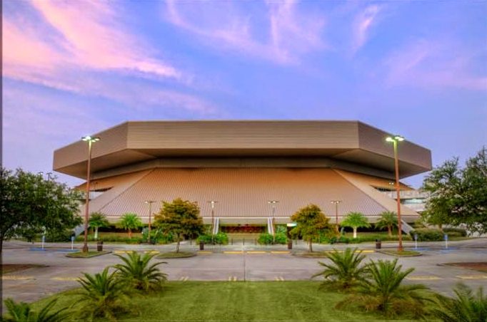 The tan, flat-topped exterior of UNO Lakefront Arena at dusk, with palm trees and lamp posts along the empty plaza in front