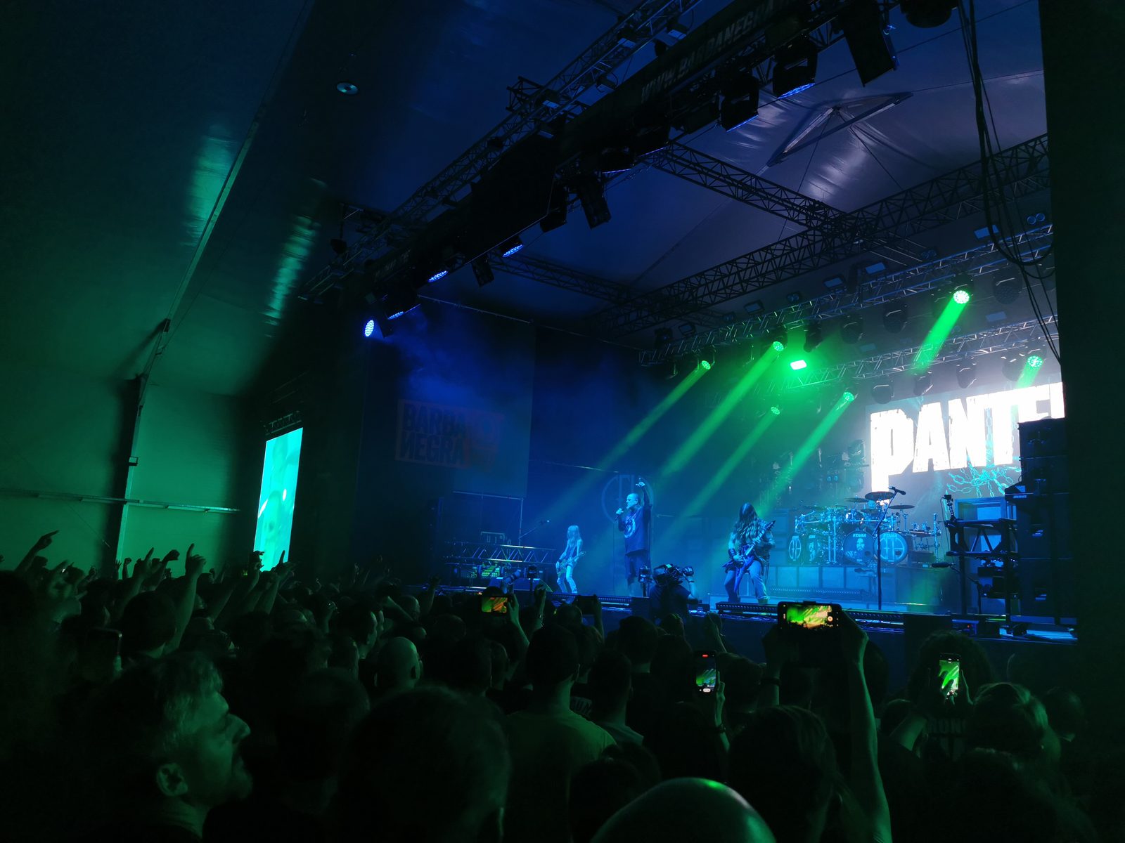 A crowd with raised hands and phones under green and blue stage lights, facing a stage with the Pantera name on a banner at the right