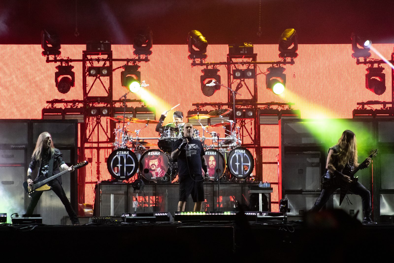 Pantera on a red-lit stage: a bassist on the left, Phil Anselmo at center in a black T-shirt and shorts holding a microphone at chest height, a guitarist on the right, and a drummer behind a kit with PH logos