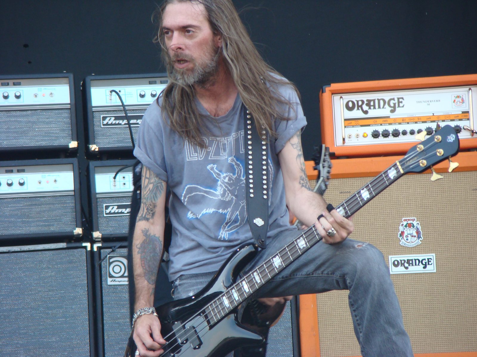 Rex Brown, long-haired and bearded in a gray Led Zeppelin T-shirt, playing a black bass in front of stacked Ampeg and orange Orange amplifiers