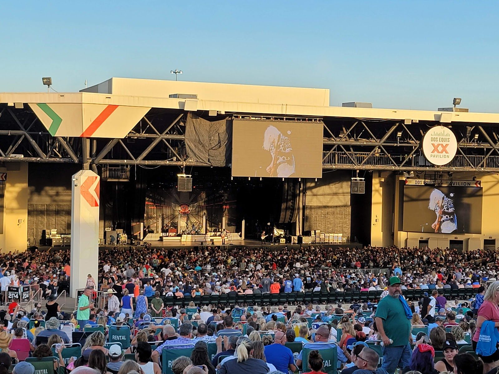 A packed Dos Equis Pavilion seen from the lawn at sunset, with a band on the covered stage, two video screens and the round Dos Equis XX Pavilion sign on the roof truss