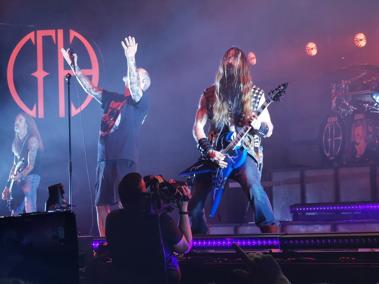 Pantera on a purple-lit stage: a long-haired bassist at left, Phil Anselmo with both arms raised in front of the red PH logo, and a long-haired guitarist playing a blue V-shaped guitar, with a camera operator in the foreground