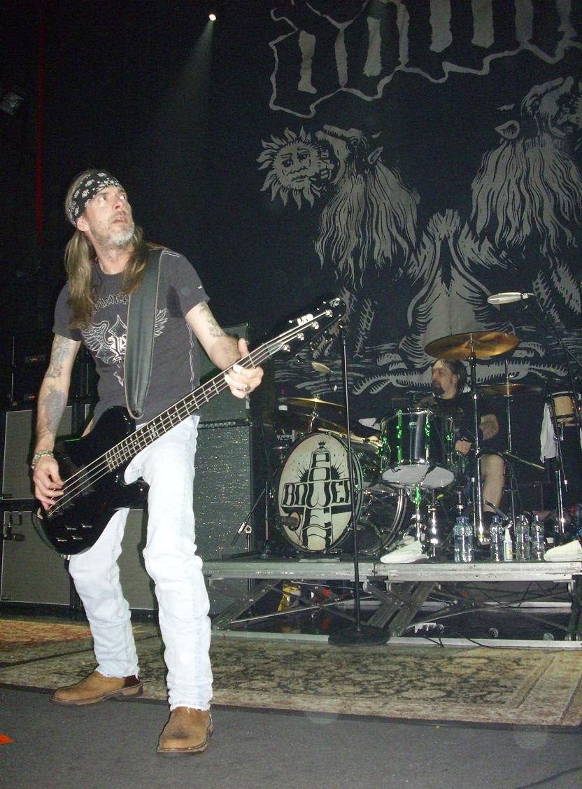 Rex Brown in a black bandana, grey T-shirt and white jeans playing a black bass in front of a drum kit and a large painted Down backdrop