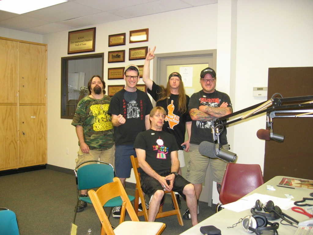 Five men posing in the KNON radio studio in Dallas beside broadcast microphones, one throwing devil horns