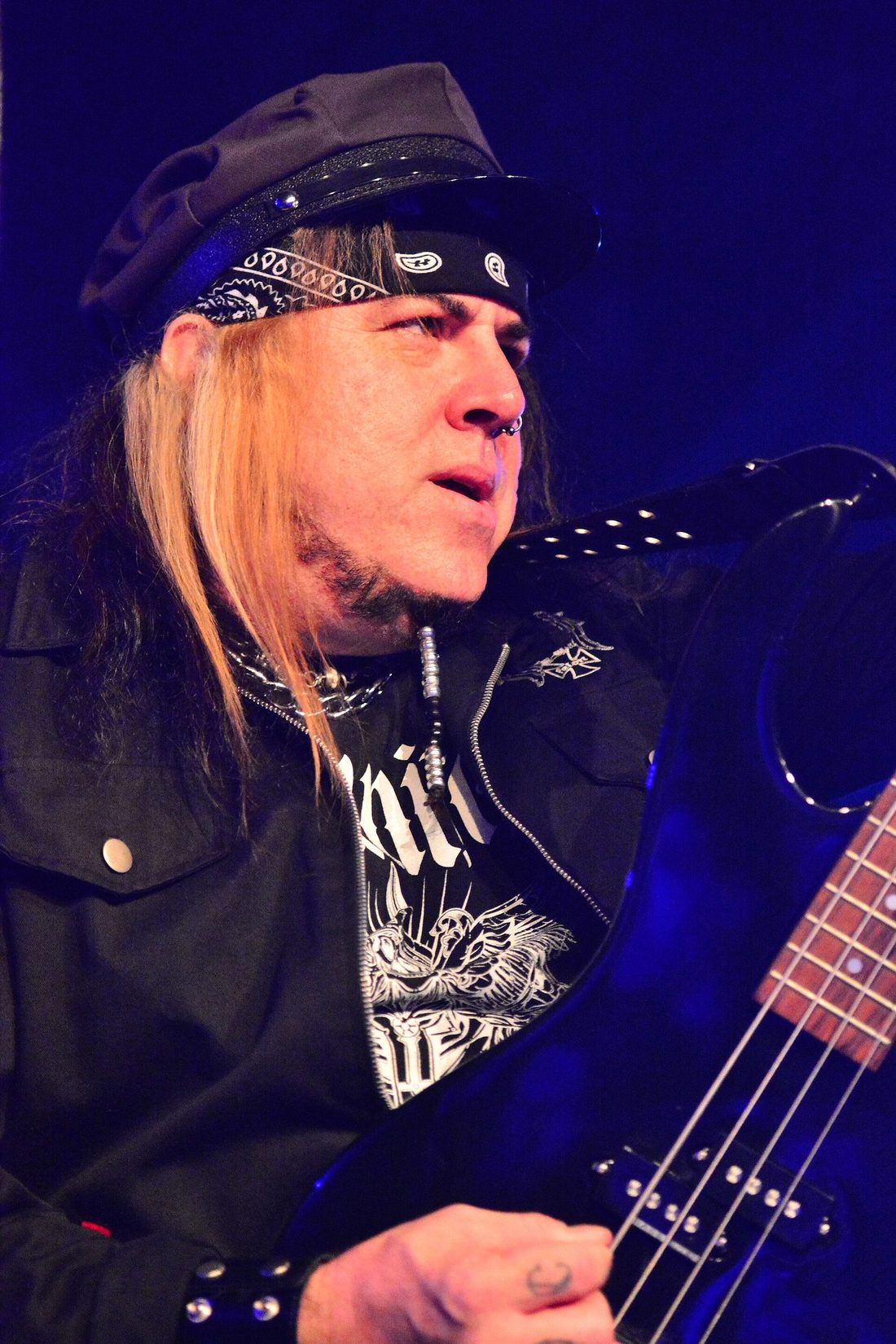 Don Van Stavern in close-up playing a black bass, long blond hair under a black captain's cap and paisley bandana, lit in deep blue