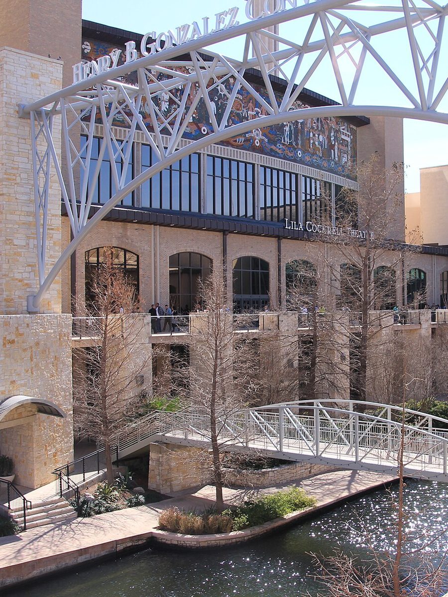 The Henry B. Gonzalez Convention Center in San Antonio, its name on a white steel arch over a limestone building with a tiled mural, above a River Walk channel and footbridge