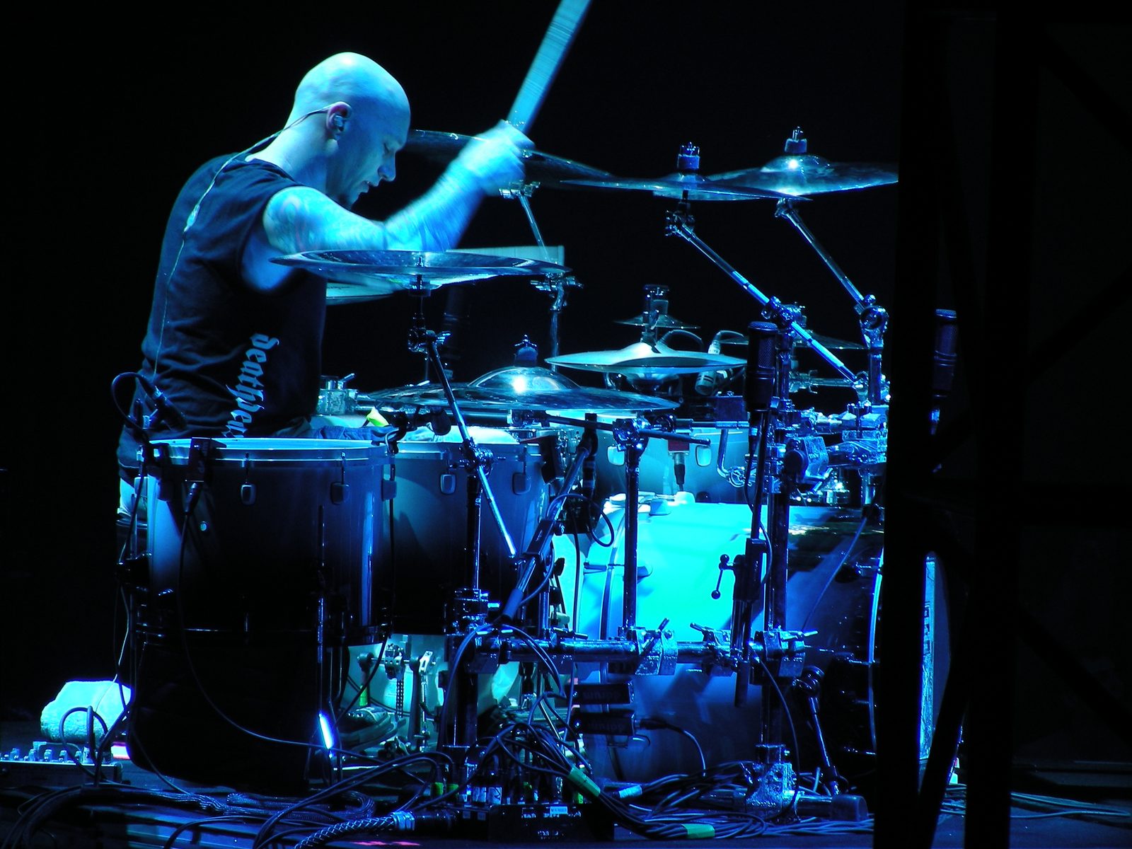 Dave McClain, shaved head and tattooed arms, drumming side-on under blue stage light, one stick raised above a large kit of toms and cymbals