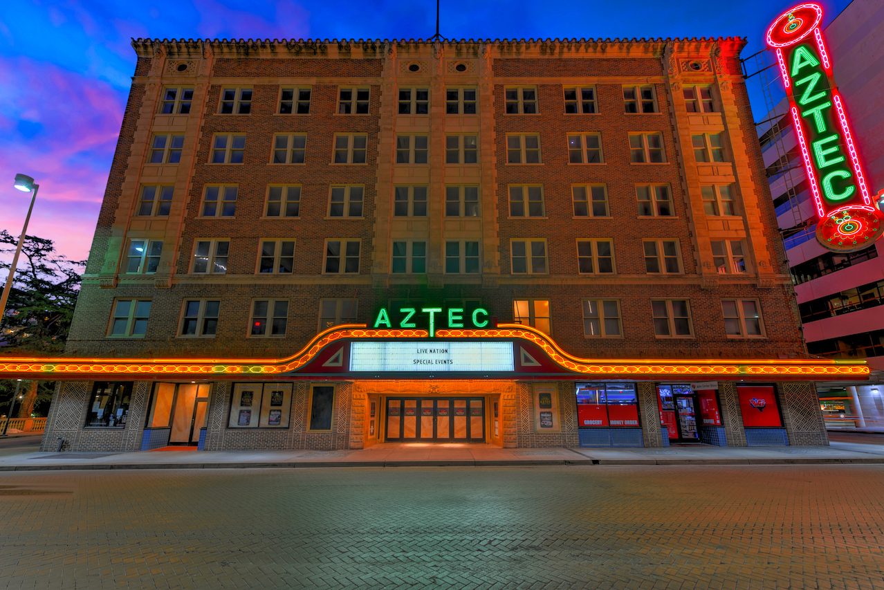 The six-story brick Aztec Theatre building at dusk with its green neon AZTEC sign, a lit marquee and a tall vertical neon blade sign at the corner