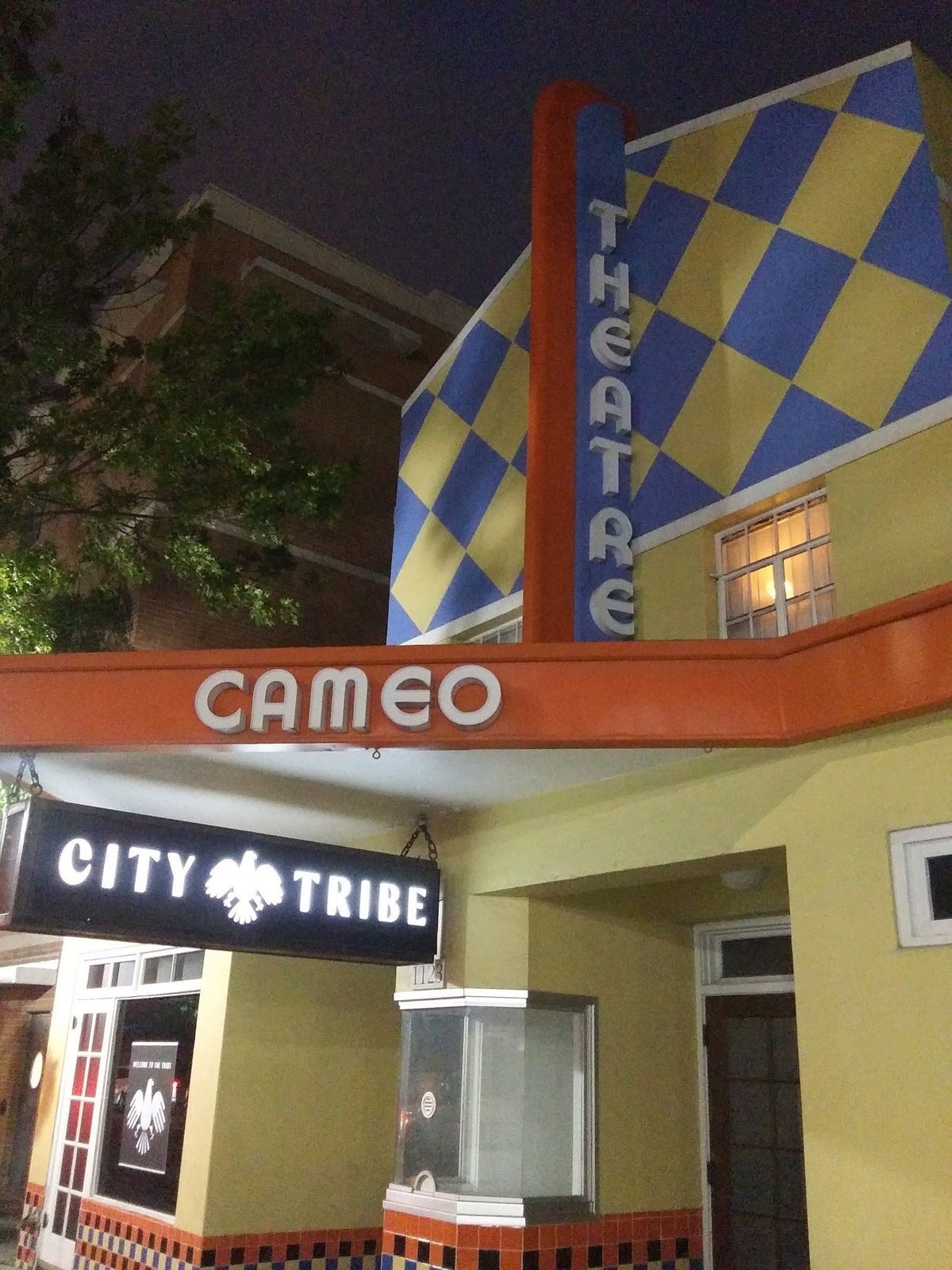 The yellow Cameo building at night, with CAMEO in white letters on an orange canopy, a vertical THEATRE sign against blue and yellow diamonds, and the street number 1123 above the old ticket booth