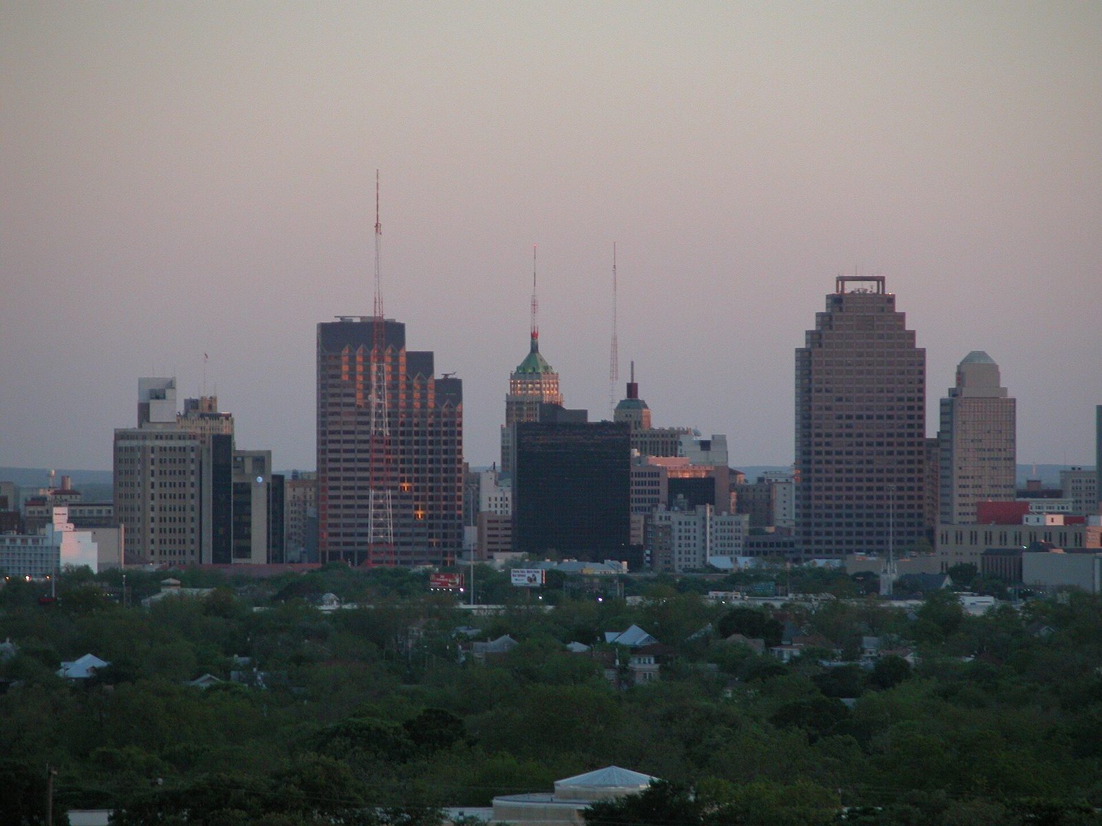 The downtown San Antonio skyline at dusk seen from the north, with the green-topped Tower Life Building and radio masts rising above a canopy of trees