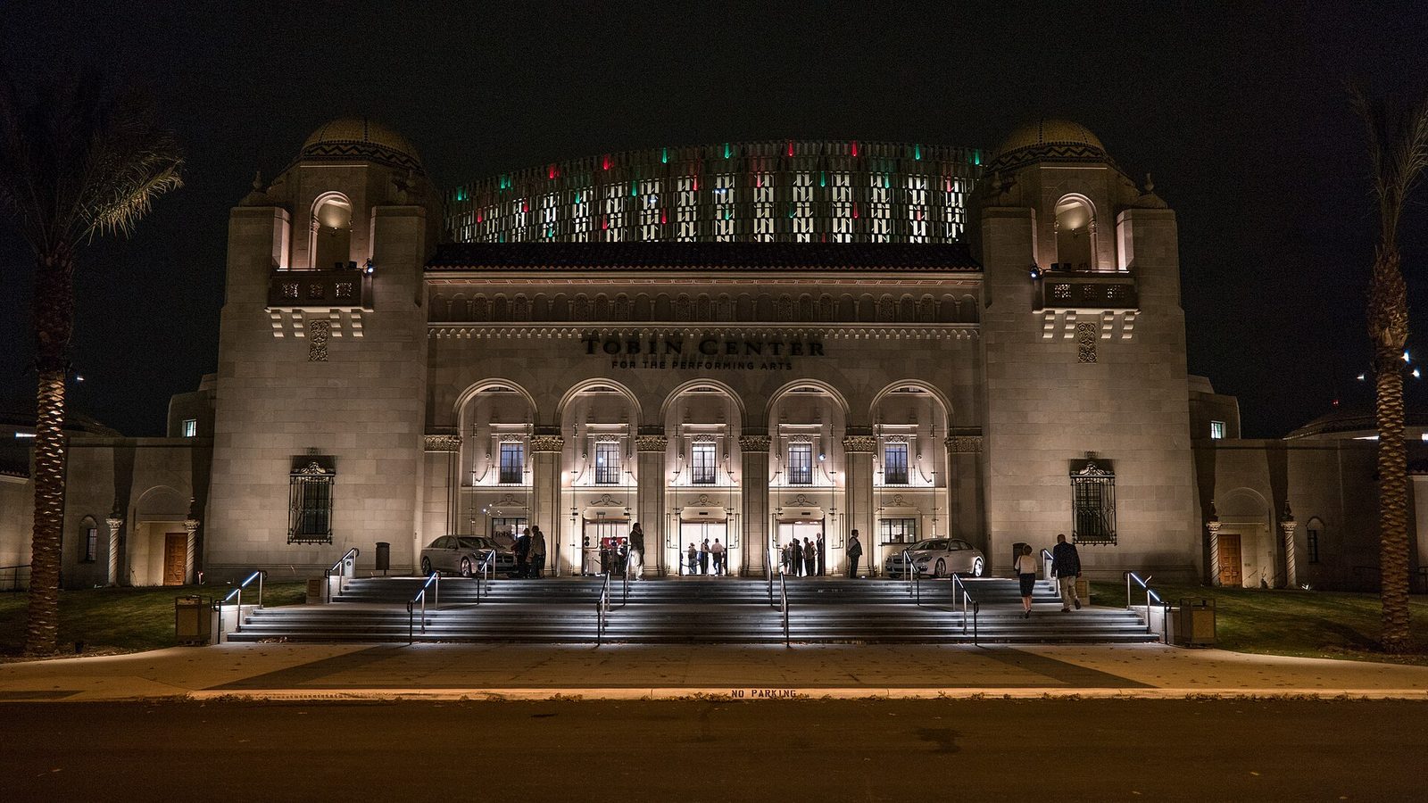 The lit Spanish Colonial Revival facade of the Tobin Center at night, with arched entrances, twin domed towers and a modern metal shell rising behind